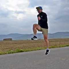 Mężczyzna w czapce i okularach ćwiczy na drodze, podnosząc kolano. Jego sprawność fizyczna jest widoczna w dynamicznym ruchu.