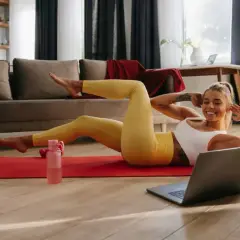 Uśmiechnięta kobieta wykonuje ćwiczenia bez sprzętu w domu, patrząc na laptopa.