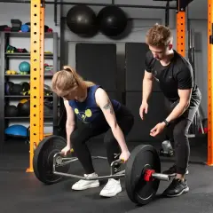 Kobieta wykonuje martwy ciąg z trap baru, trener koryguje jej pozycję. Ćwiczenie angażuje wiele mięśni.