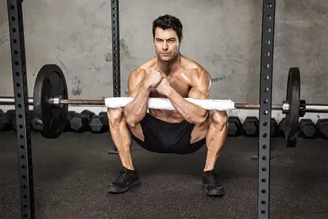 Mężczyzna wykonuje ćwiczenie zercher squat z gryfem opartym na przedramionach.