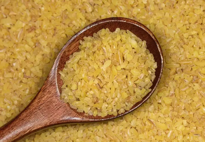 Drewniana łyżka pełna drobnej, żółtej kaszy bulgur na tle rozsypanych ziaren. To zdrowy składnik wielu dań.