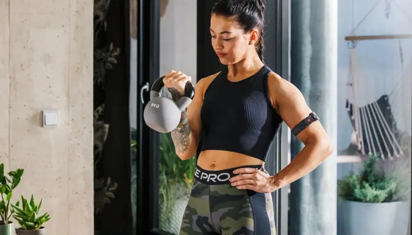 Kobieta ćwiczy z kettlebell, co przyspiesza metabolizm. Na ramieniu ma opaskę monitorującą trening.