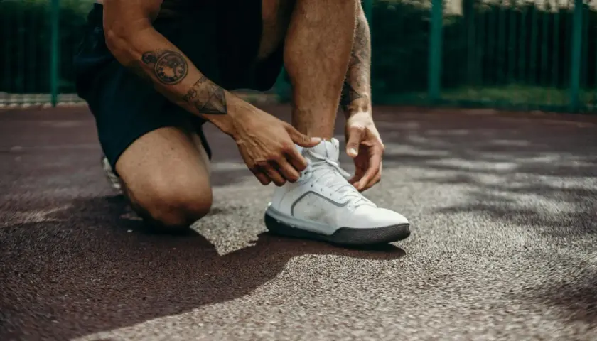 Mężczyzna z tatuażami na ramieniu klęczy, sznurując białe buty sportowe. Może to być przygotowanie do treningu lub ulga po bólu kolan po ćwiczeniach.