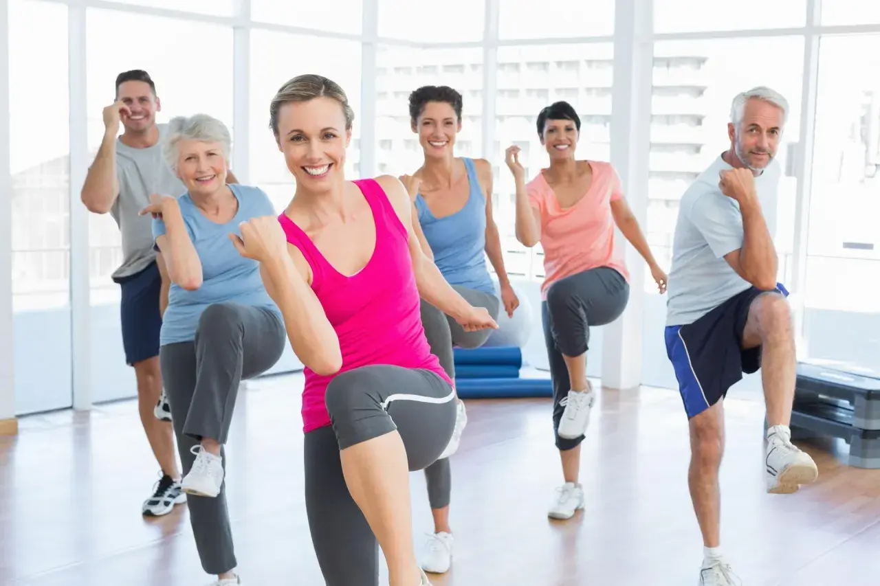 Uśmiechnięci ludzie w różnym wieku ćwiczą w sali gimnastycznej, wykonując ruchy inspirowane **les mills bodybalance**.