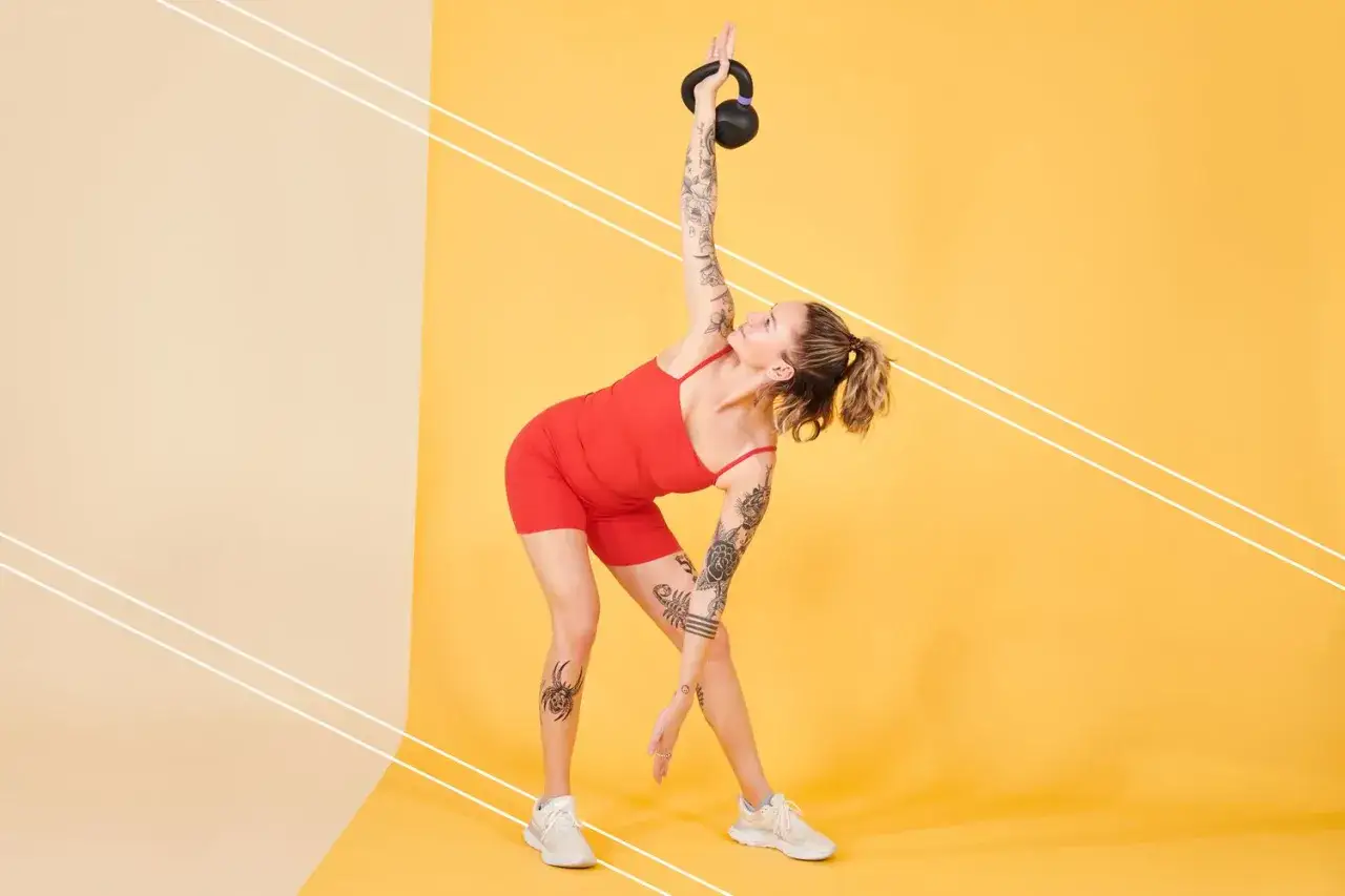 Kobieta w czerwonym stroju wykonuje ćwiczenia z kettlebell, unosząc go nad głowę.